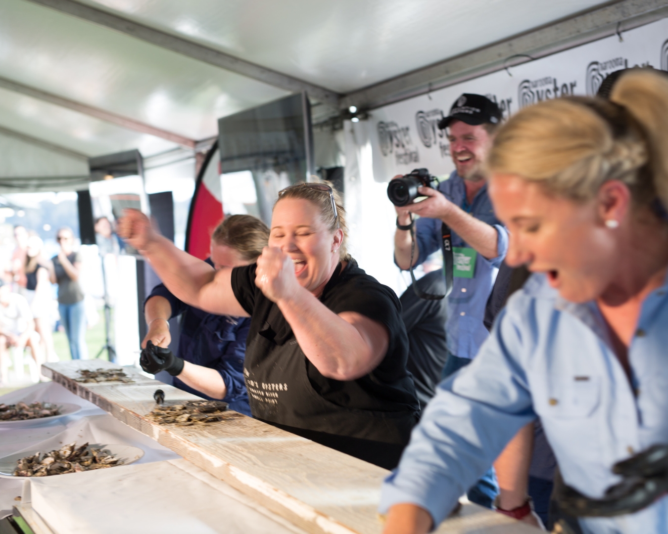 Three women in a race to shuck oysters. The woman in the middle has finished and raised her hands in victory.