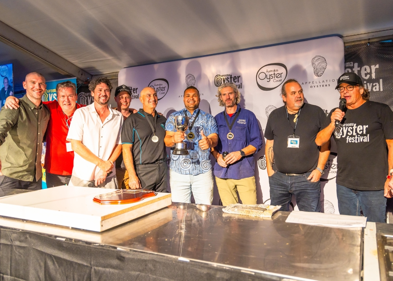 A group of men stand on stage surrounding Gerard Denis, winner of the 2023 Shucking Competition and inaugural winner of the John Susman trophy