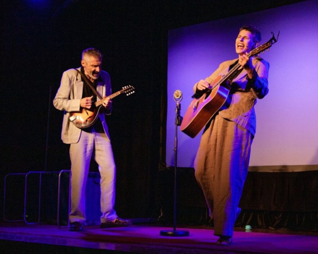 Deborah Conway & Willy Zygier are performing on stage, Deb is singing and playing guitar and Willy is playing guitar.
