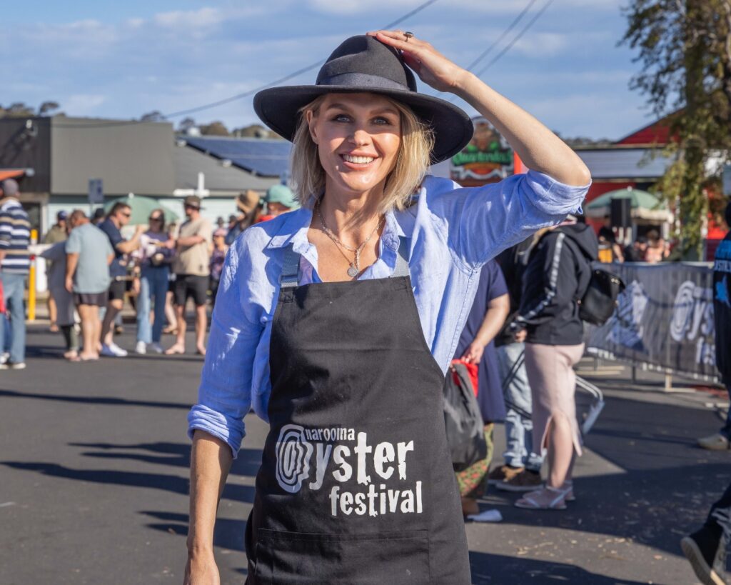 a woman with blond hair and wearing a blue shirt, black apron and hat is standing in a festival crowd and smiling at the camera