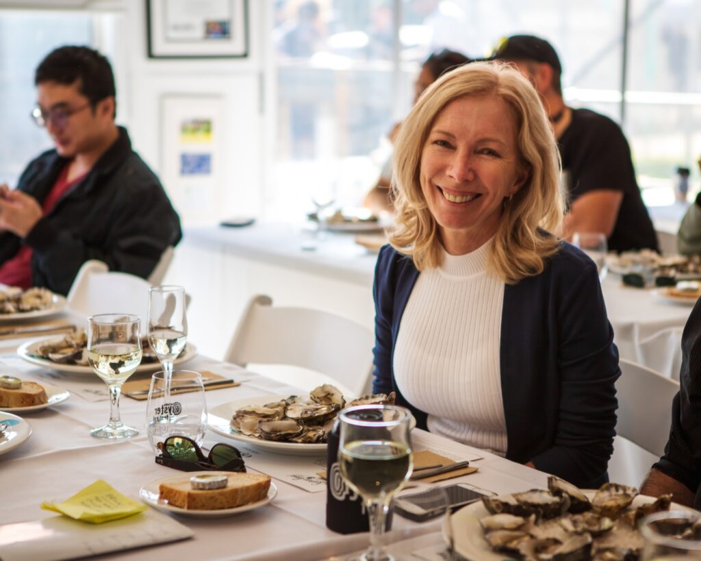 A woman sitd at a table with a plate of oysters in front of her and a glass of wine. she is delighted.