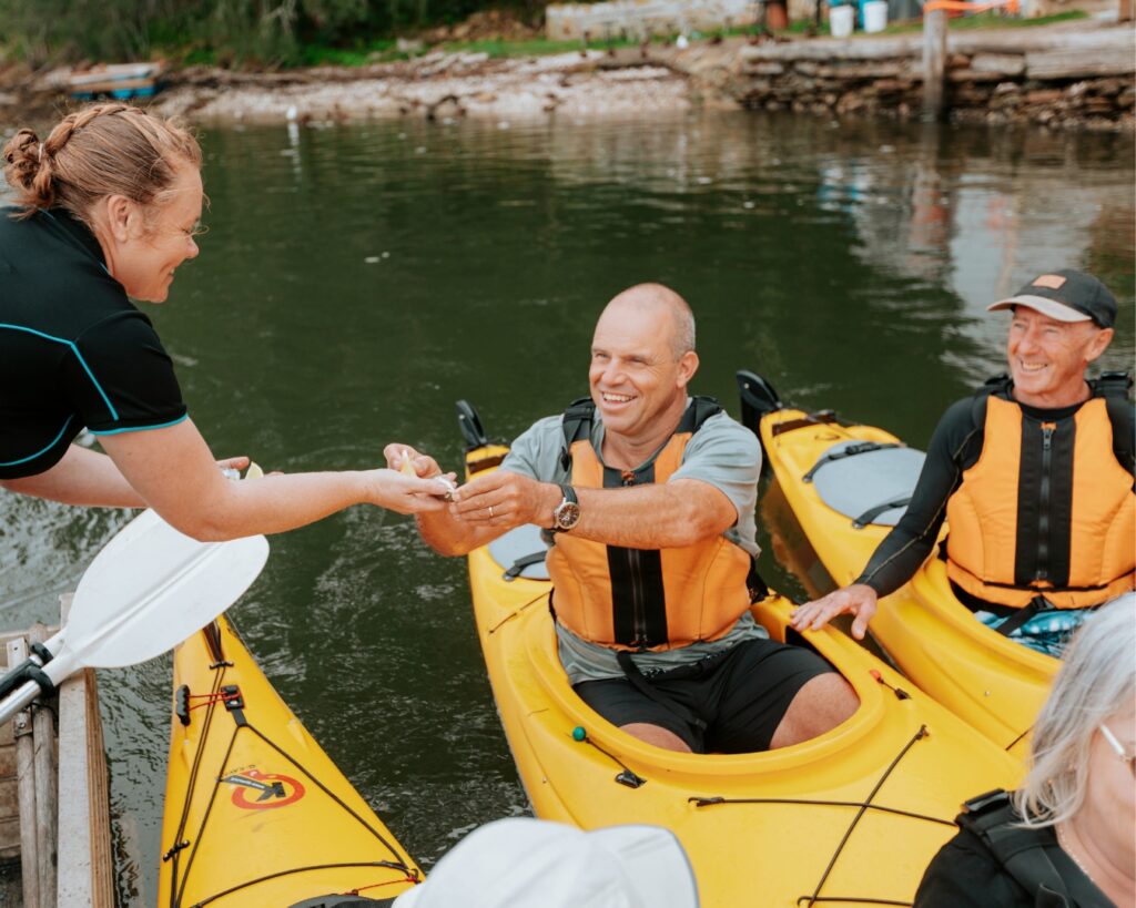 Oyster farmer Jady Norris hands freshly shucked rock oysters to a man sitting in a kayak, he is delighted!